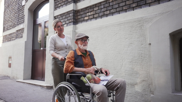 Granddaughter pushing senior man in wheelchair on street. Female caregiver and elderly man enjoying a warm autumn day, going home from shopping.