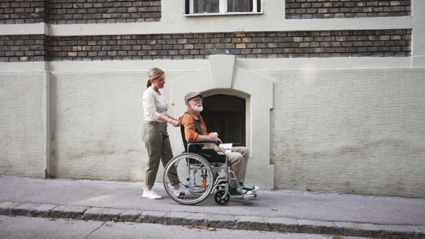 Granddaughter pushing senior man in wheelchair on street. Female caregiver and elderly man enjoying a warm autumn day, going home from shopping.