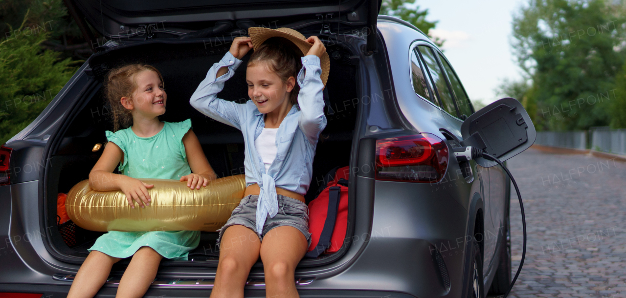 Cute little girls sitting in a boot while waiting for charging car before travelling on summer holiday.