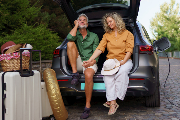 A middle-aged couple sitting in trunk while waiting for charging car before travelling on summer holiday.