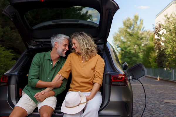 A middle-aged couple sitting in trunk while waiting for charging car before travelling on summer holiday.