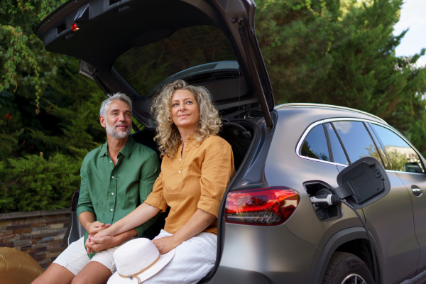 A middle-aged couple sitting in trunk while waiting for charging car before travelling on summer holiday.