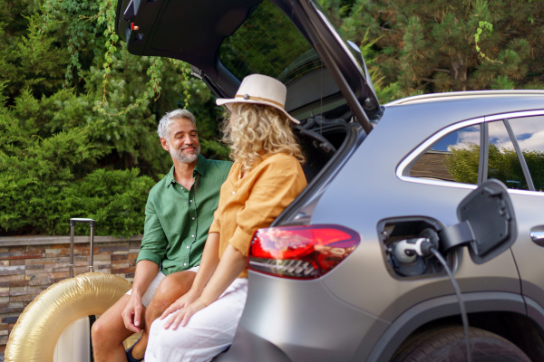 A middle-aged couple sitting in trunk while waiting for charging car before travelling on summer holiday.