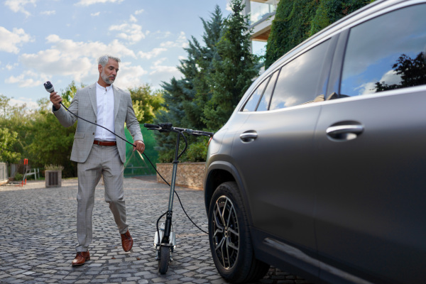 Businessman in suit on way to work with eletric scooter and charging his electric car. Concept of eco commuting and green transport.