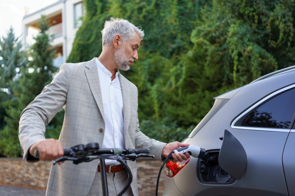 Close up of businessman in suit on way to work standing at an eletric scooter and charging his electric car. Concept of eco commuting and green transportation.