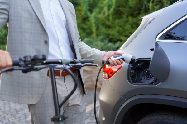 Close up of businessman in suit on way to work standing at an eletric scooter and charging his electric car. Concept of eco commuting and green transportation.