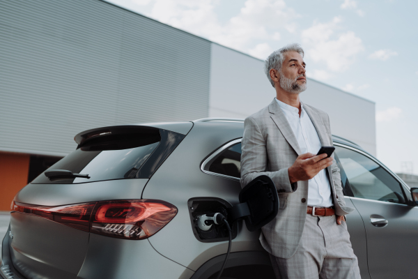 A businessman holding smartphone while charging car at electric vehicle charging station, closeup.