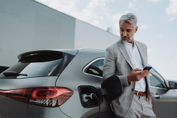 A businessman holding smartphone while charging car at electric vehicle charging station, closeup.