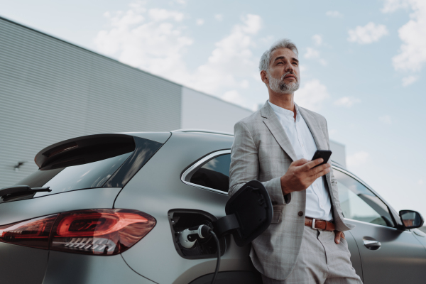 A businessman holding smartphone while charging car at electric vehicle charging station, closeup.