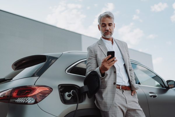 A businessman holding smartphone while charging car at electric vehicle charging station, closeup.