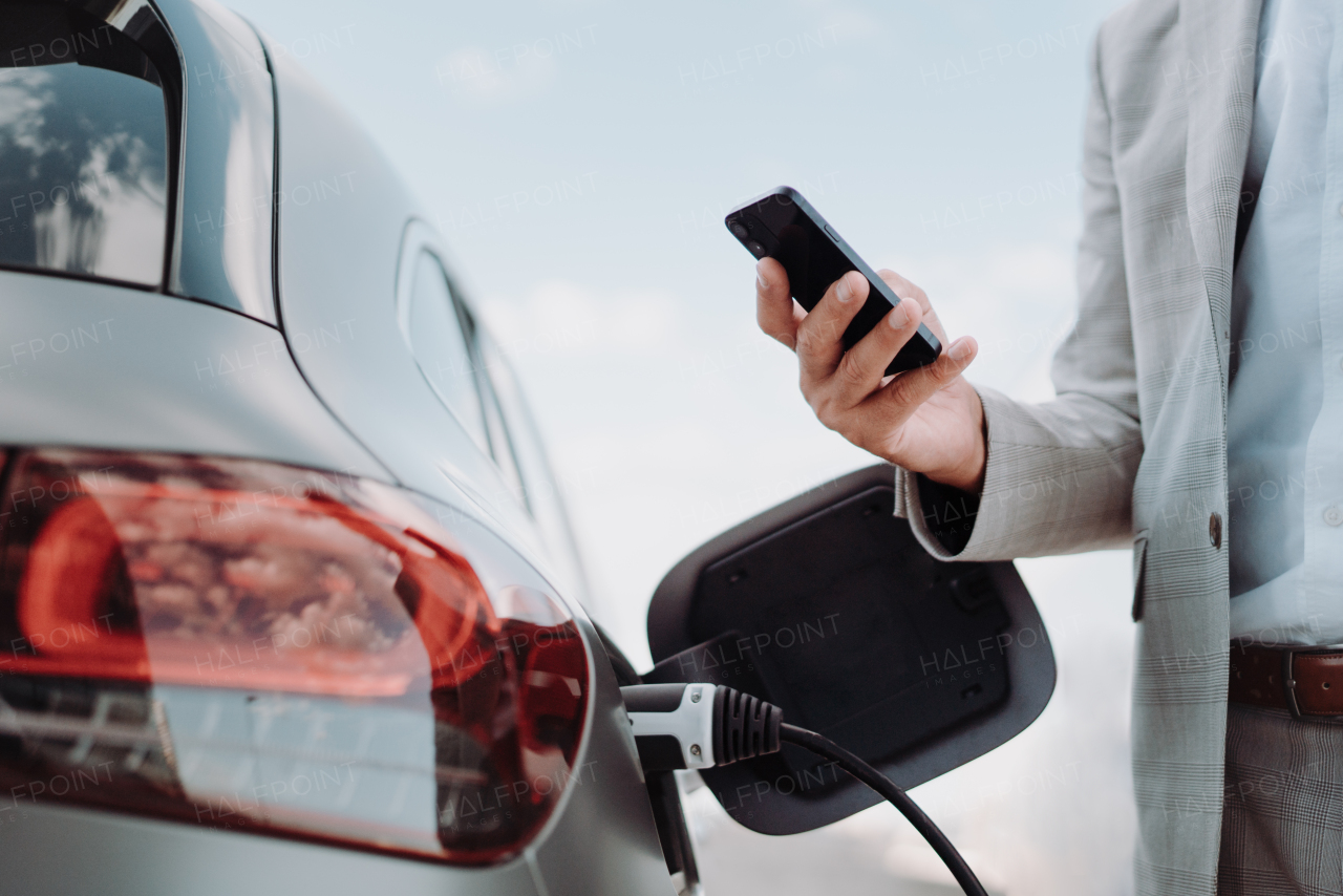 A man holding smartphone while charging car at electric vehicle charging station, closeup.