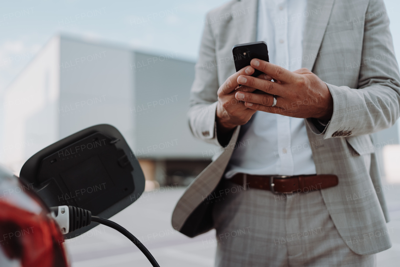 A man holding smartphone while charging car at electric vehicle charging station, closeup.