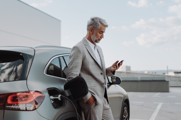 A businessman holding smartphone while charging car at electric vehicle charging station, closeup.