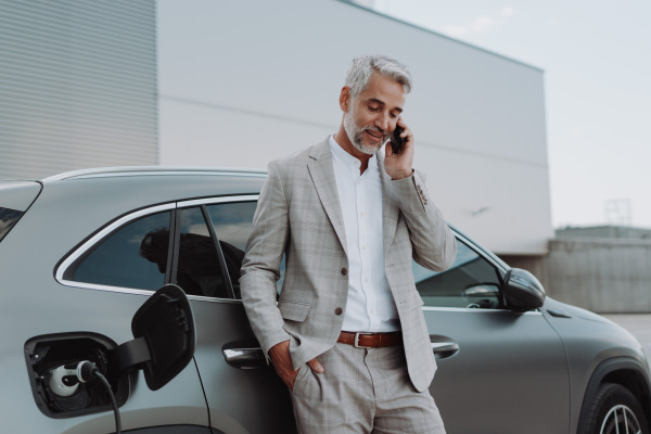 A businessman holding smartphone while charging car at electric vehicle charging station, closeup.