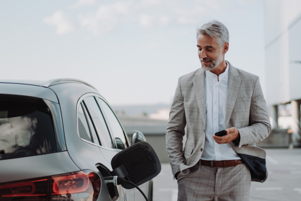 A businessman holding smartphone while charging car at electric vehicle charging station, closeup.