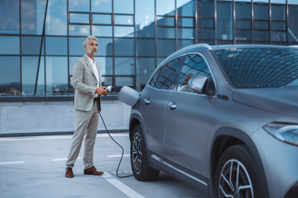 A businessman holding power supply cable at electric vehicle charging station.