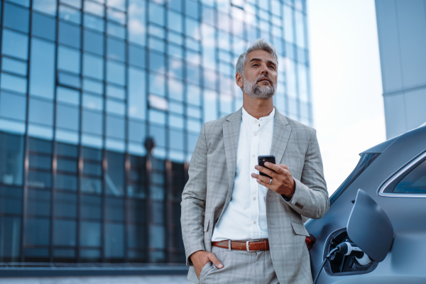 A businessman holding smartphone while charging car at electric vehicle charging station, closeup.