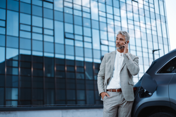 A businessman holding smartphone while charging car at electric vehicle charging station, closeup.