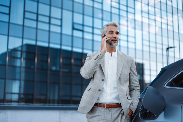 A businessman phoneing while charging car at electric vehicle charging station, closeup.