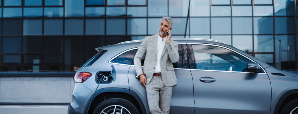 A businessman holding smartphone while charging car at electric vehicle charging station, closeup.