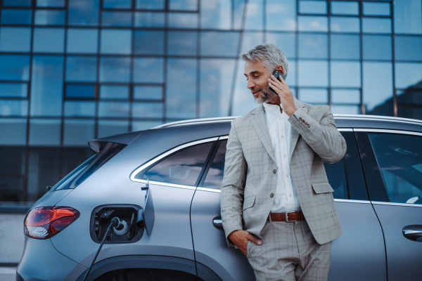 A businessman holding smartphone while charging car at electric vehicle charging station, closeup.