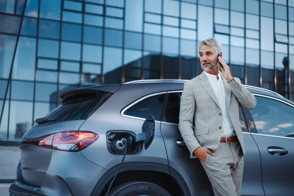 A businessman phoneing while charging car at electric vehicle charging station, closeup.