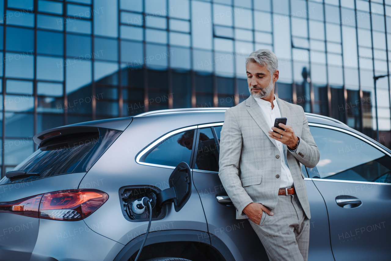 A businessman holding smartphone while charging car at electric vehicle charging station, closeup.