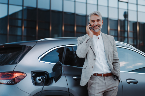 A businessman holding smartphone while charging car at electric vehicle charging station, closeup.