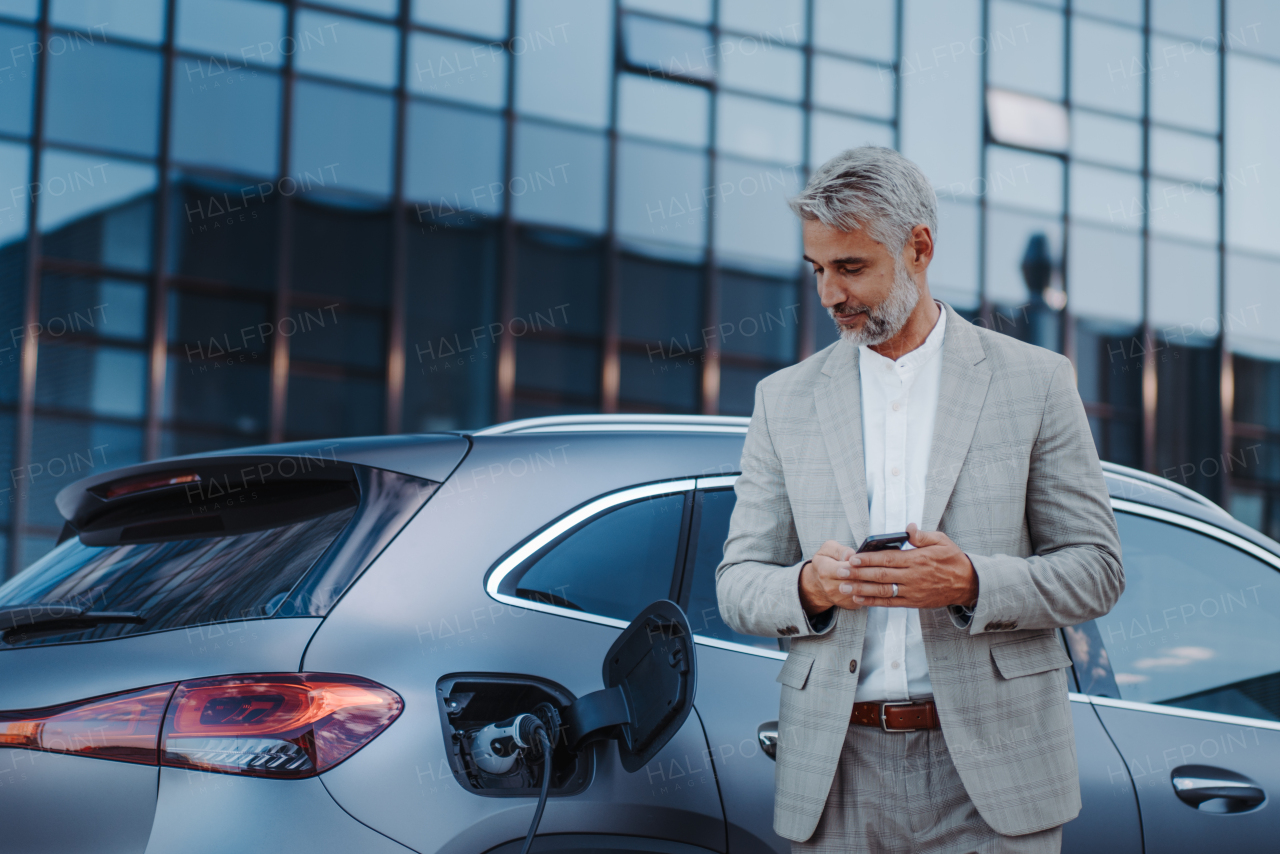 A businessman holding smartphone while charging car at electric vehicle charging station, closeup.