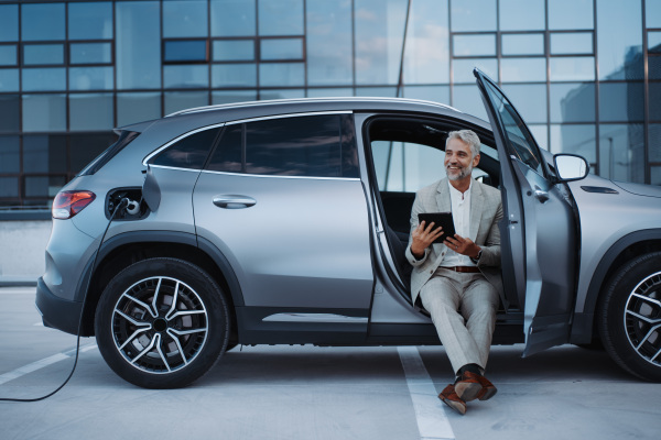 Businessman using tablet while charging a car at electric vehicle charging station, close-up.