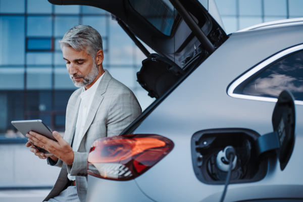 Businessman using tablet while charging a car at electric vehicle charging station, close-up.