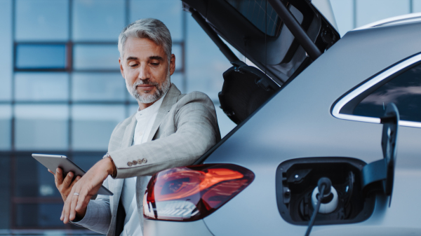 Businessman using tablet while charging a car at electric vehicle charging station, close-up.