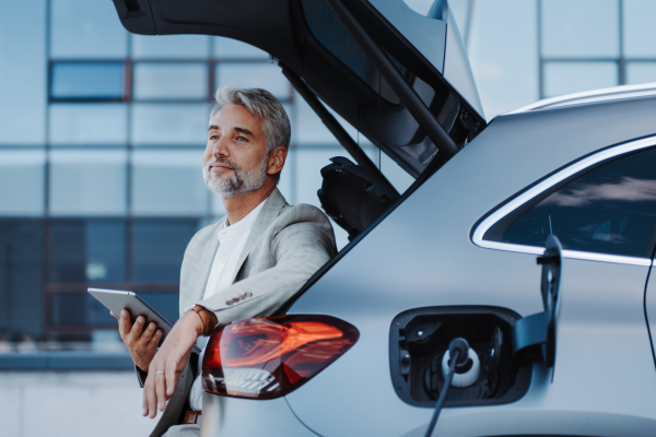 Businessman using tablet while charging a car at electric vehicle charging station, close-up.