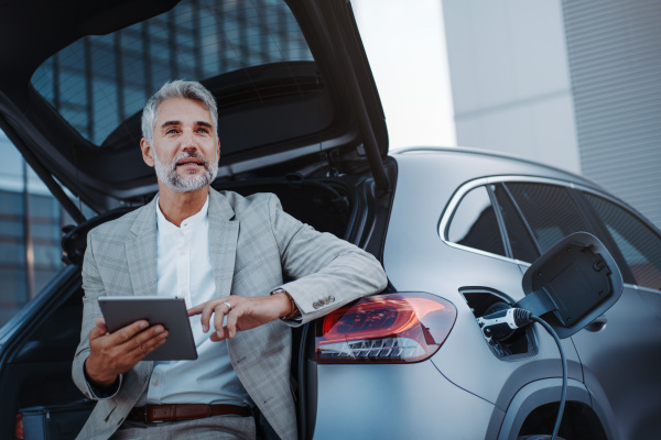 A businessman using tablet while charging car at electric vehicle charging station, closeup.