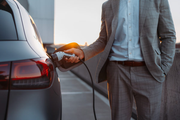 Businessman holding power supply, charging his electric car during sunset. Concept of ecology tranport.