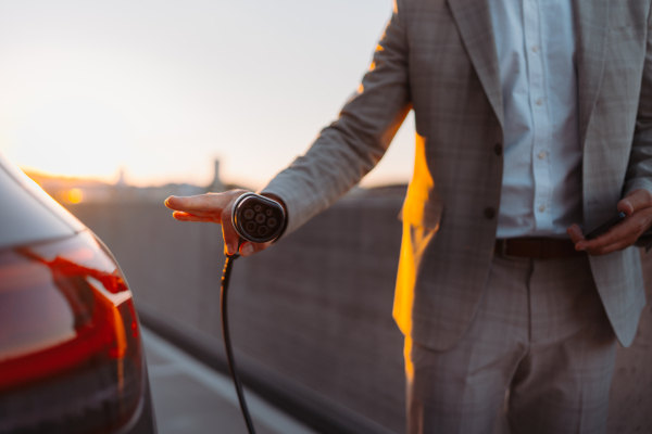 Businessman holding power supply, charging his electric car during sunset. Concept of ecology tranport.