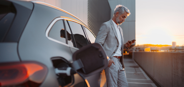 A man holding smartphone while charging car at electric vehicle charging station, closeup.