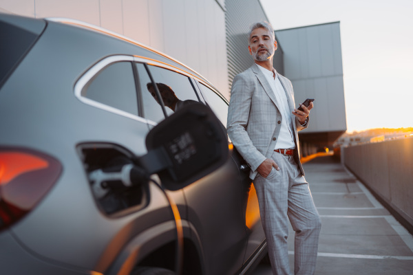 A businessman holding smartphone while charging car at electric vehicle charging station, closeup.