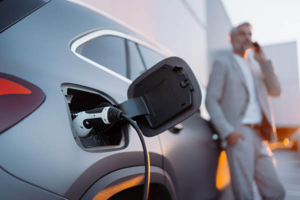 A businessman phoneing while charging car at electric vehicle charging station, closeup.