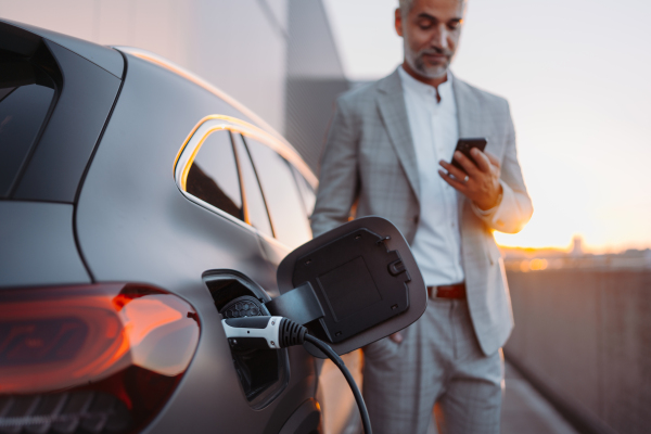 A businessman holding smartphone while charging car at electric vehicle charging station, closeup.