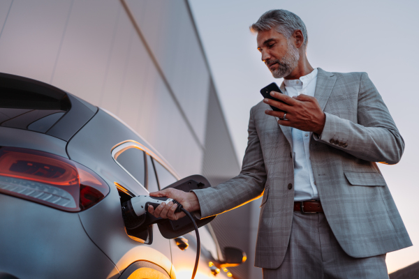 A man holding smartphone while charging car at electric vehicle charging station, closeup.