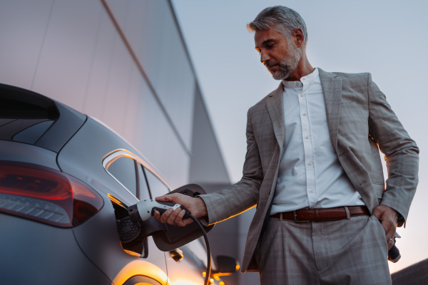 Businessman holding power supply, charging his electric car during sunset. Concept of ecology tranport.