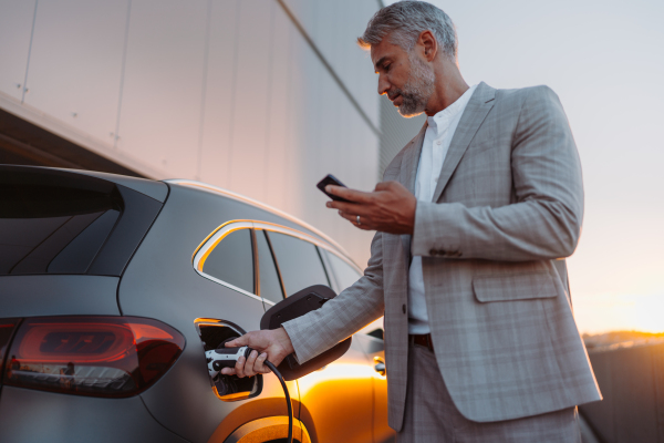 A businessman holding smartphone while charging car at electric vehicle charging station, closeup.