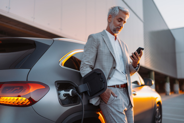 A businessman holding smartphone while charging car at electric vehicle charging station, closeup.