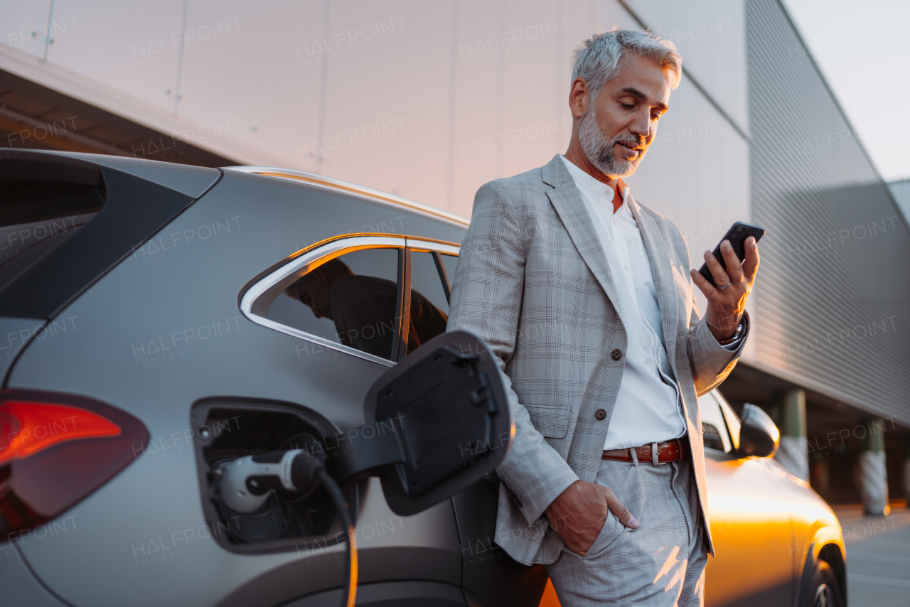 A businessman holding smartphone while charging car at electric vehicle charging station, closeup.