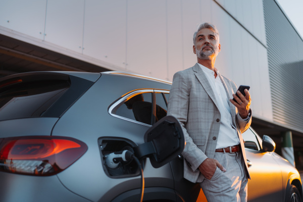 A businessman holding smartphone while charging car at electric vehicle charging station, closeup.