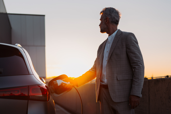 Businessman holding power supply, charging his electric car during sunset. Concept of ecology tranport.