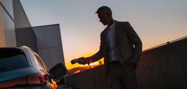 Businessman holding power supply, charging his electric car during sunset. Concept of ecology tranport.