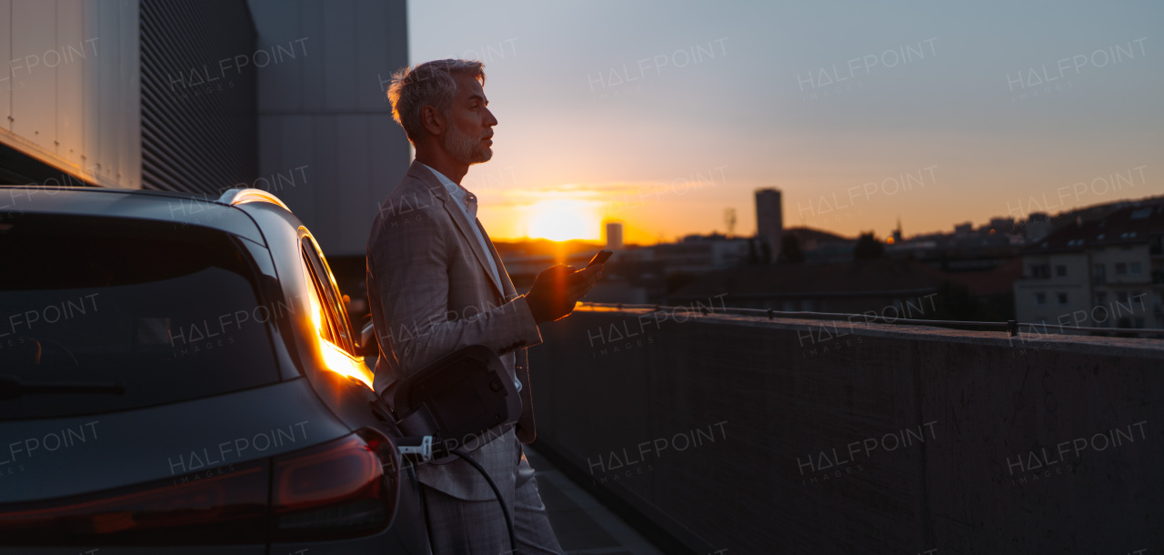 A businessman holding smartphone while charging car at electric vehicle charging station, closeup.