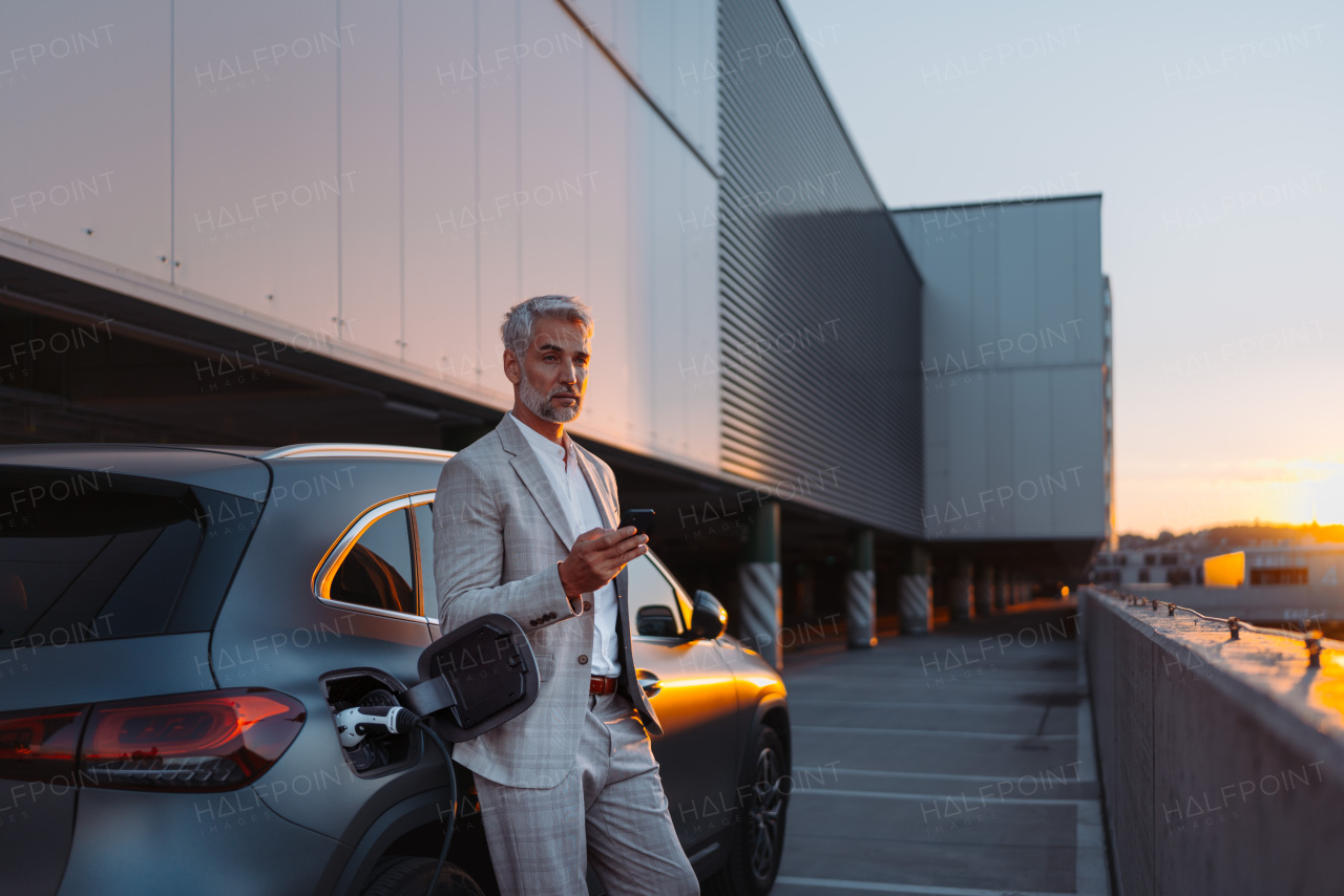 A man holding smartphone while charging car at electric vehicle charging station, closeup.